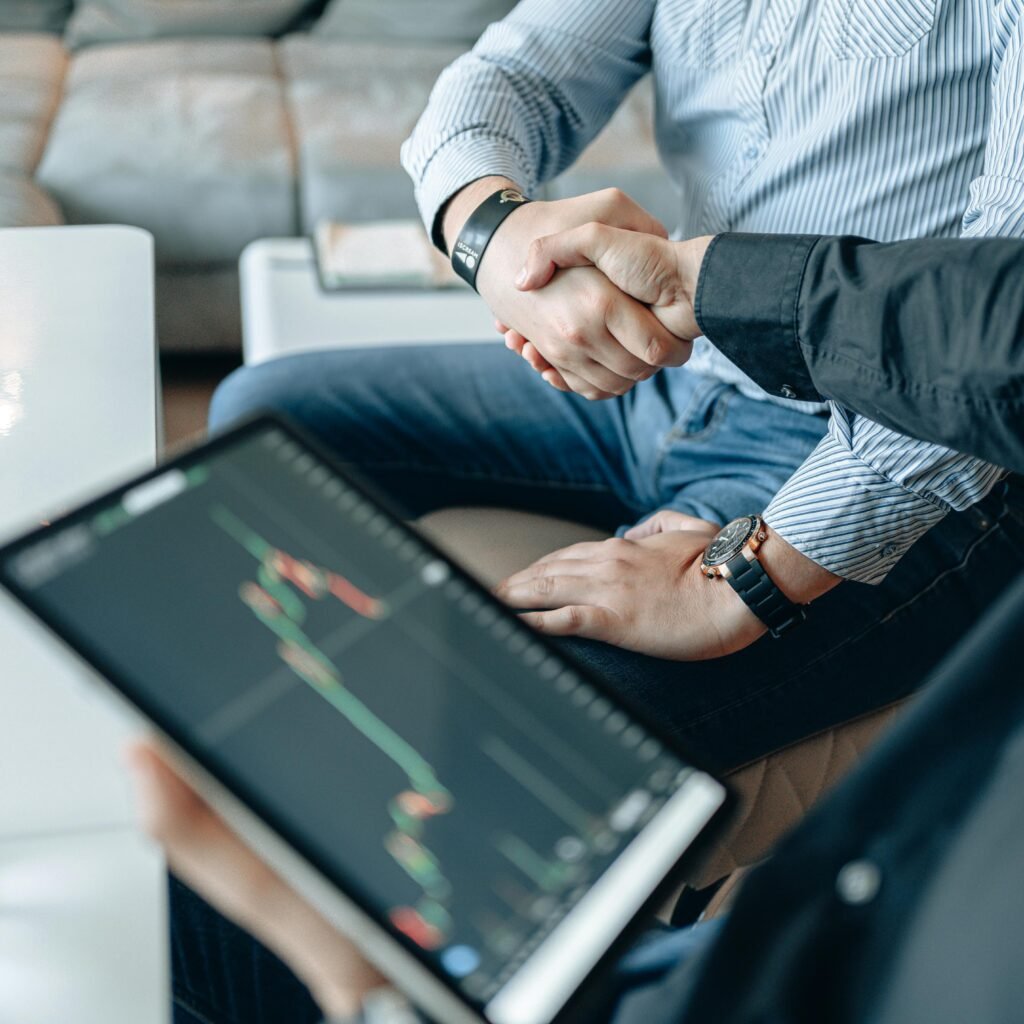 Two businesspeople shaking hands over stock market data on a tablet.