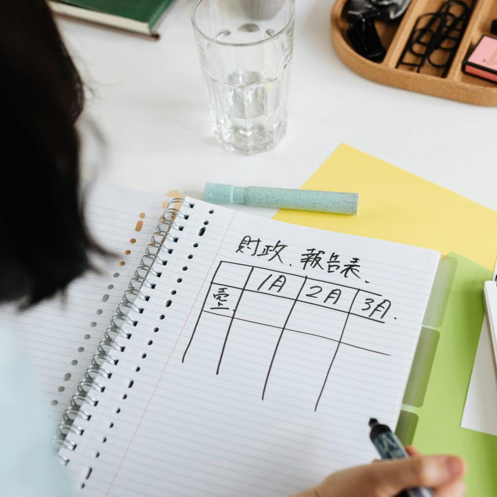 Person writing a table on a spiral notebook with planner and stationery on a desk.
