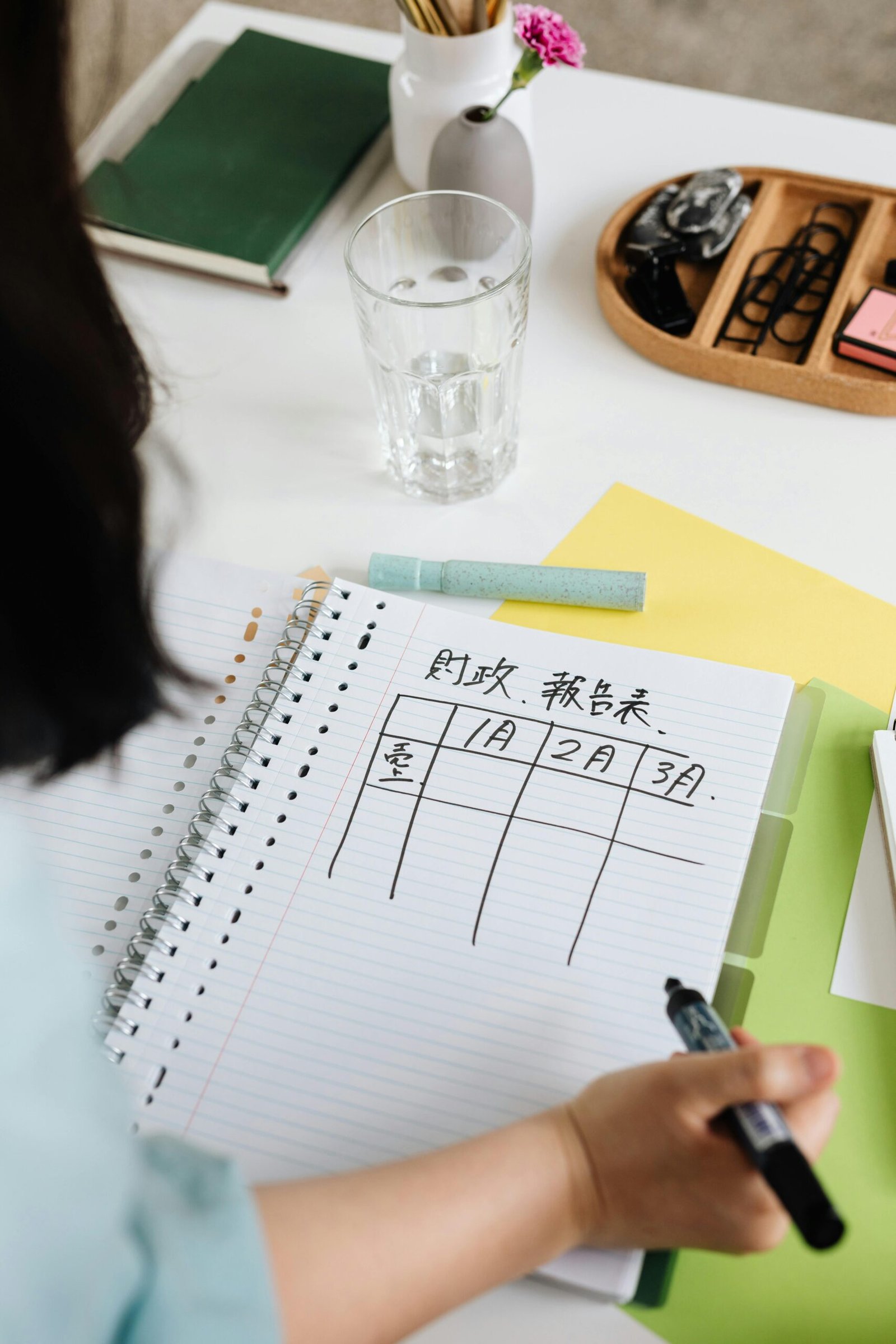 Person writing a table on a spiral notebook with planner and stationery on a desk.