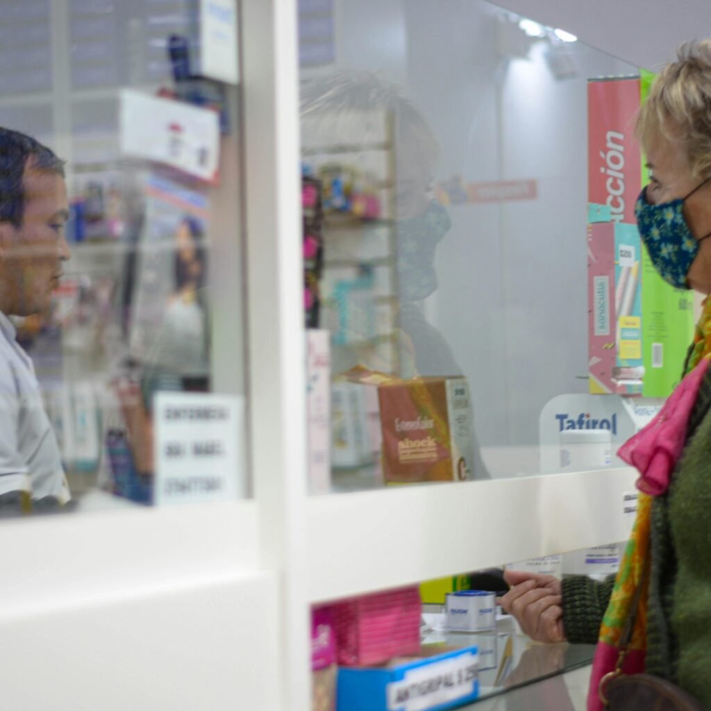 A woman with a mask speaks to a pharmacist behind a counter in a pharmacy.
