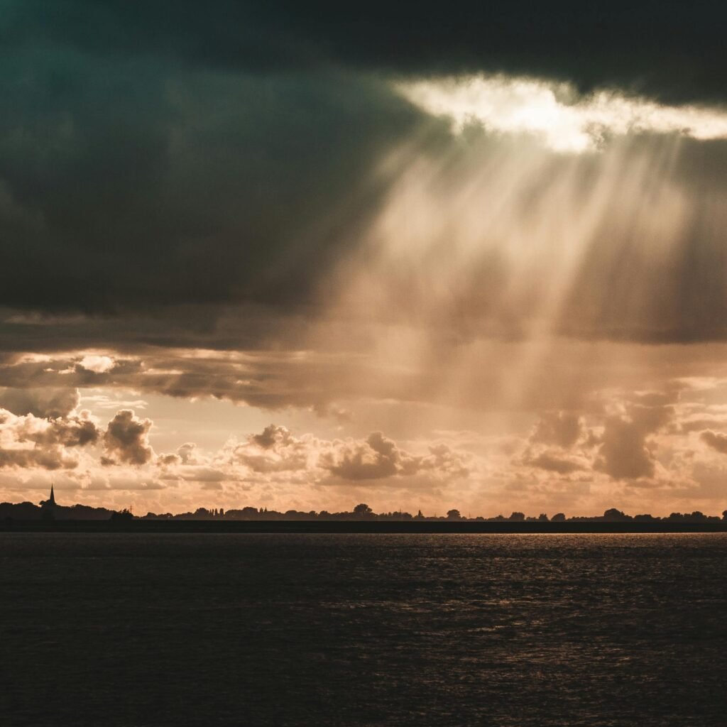 Sunbeams illuminate a serene landscape beneath dark storm clouds over a calm sea.