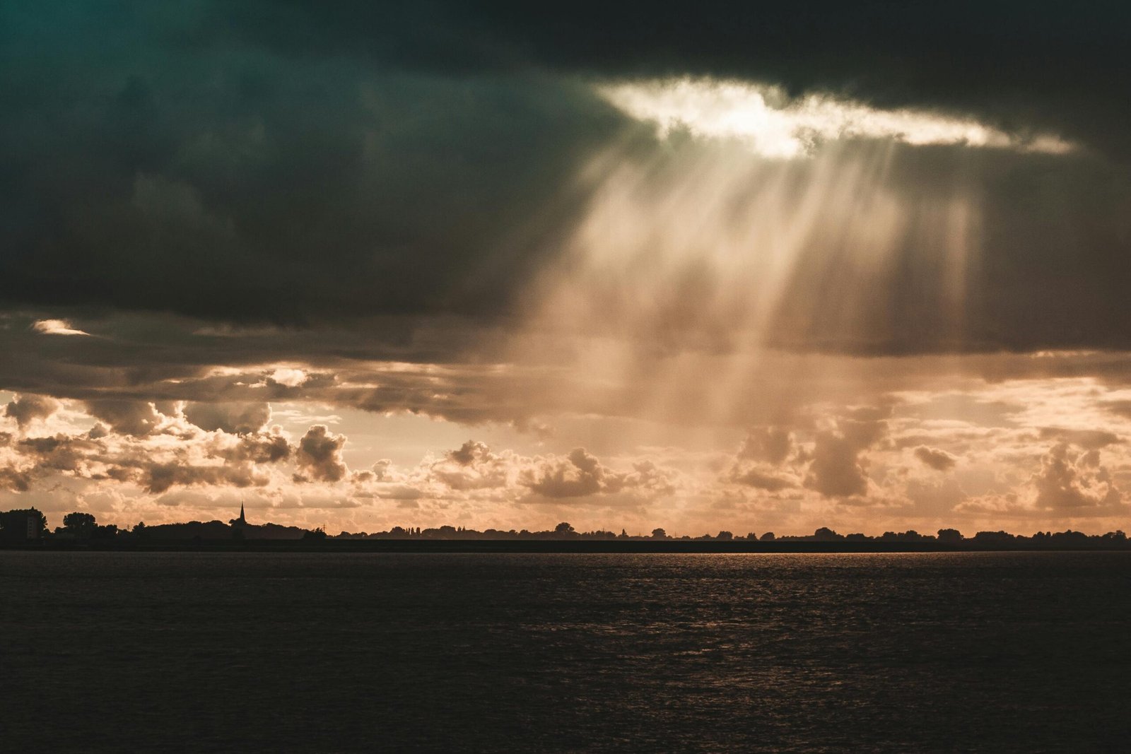 Sunbeams illuminate a serene landscape beneath dark storm clouds over a calm sea.