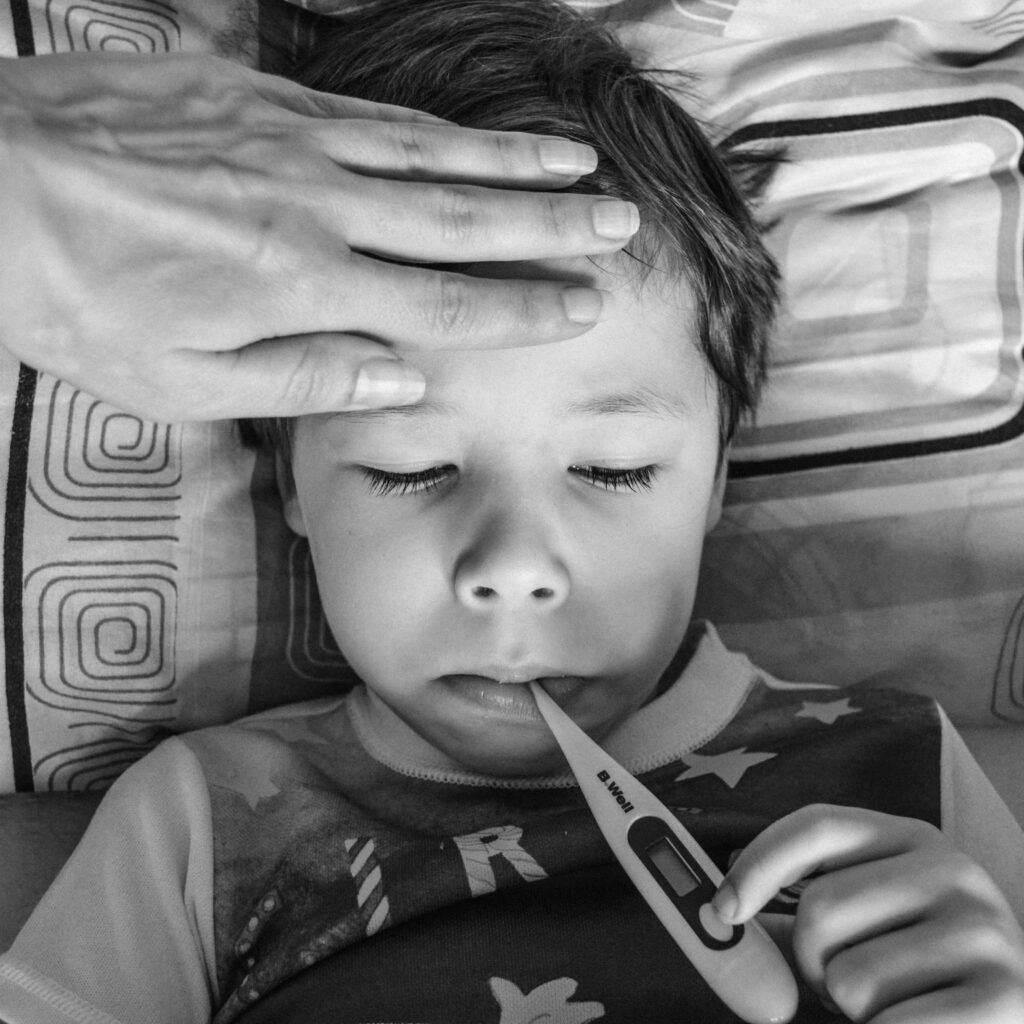 Black and white from above of unrecognizable person touching forehead of unemotional child in casual clothing holding electronic thermometer in mouth