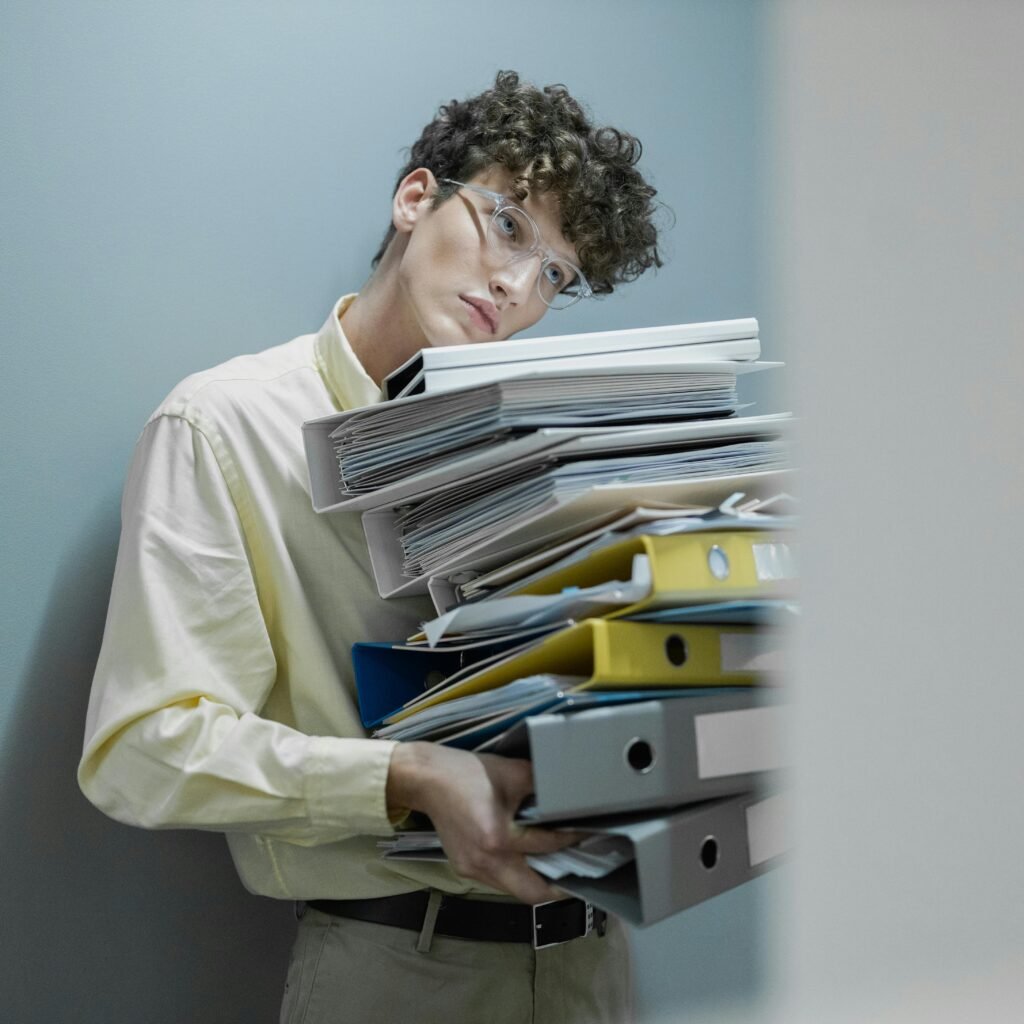 Young man overwhelmed with a stack of folders symbolizes workplace stress.