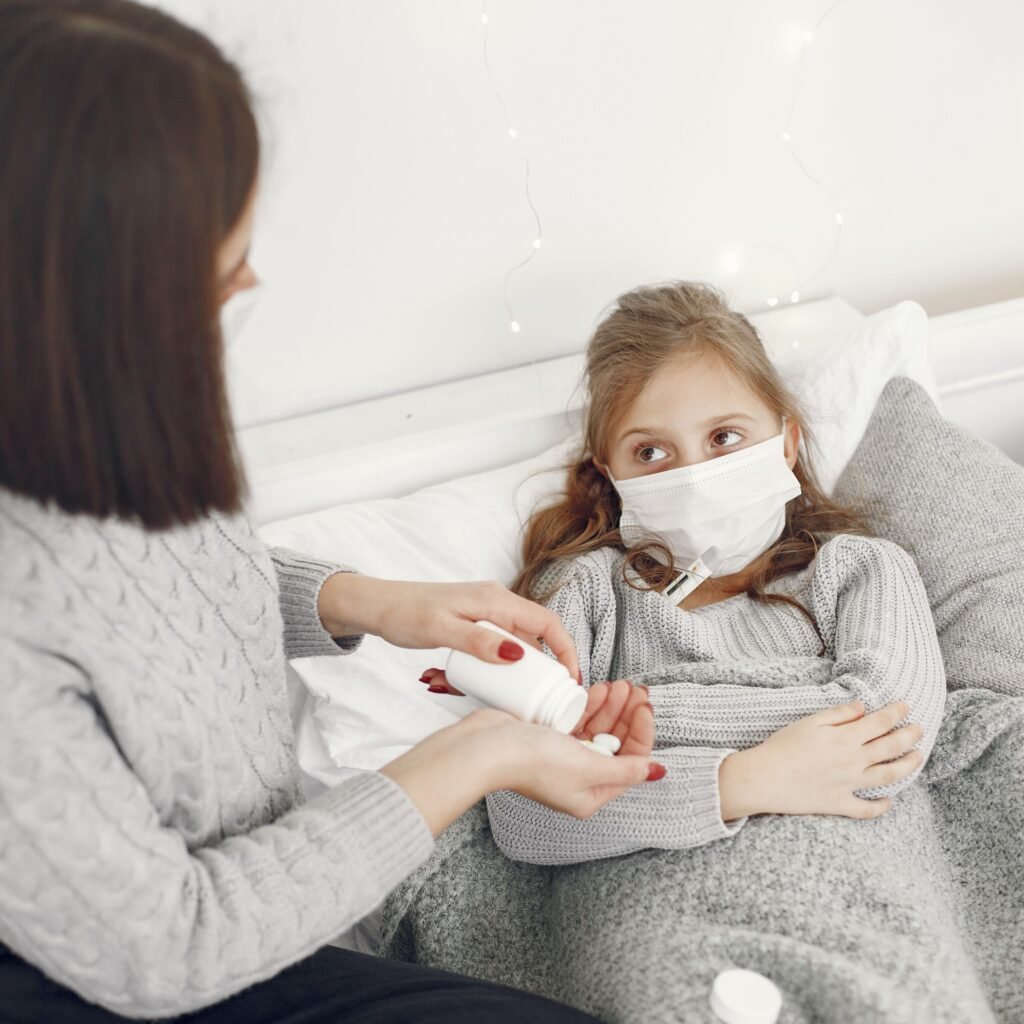 A mother helps her sick daughter wearing a face mask in bed, offering medicine.