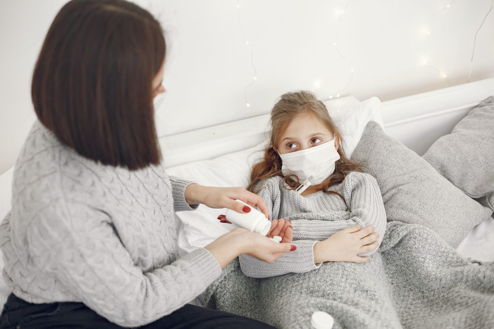 A mother helps her sick daughter wearing a face mask in bed, offering medicine.