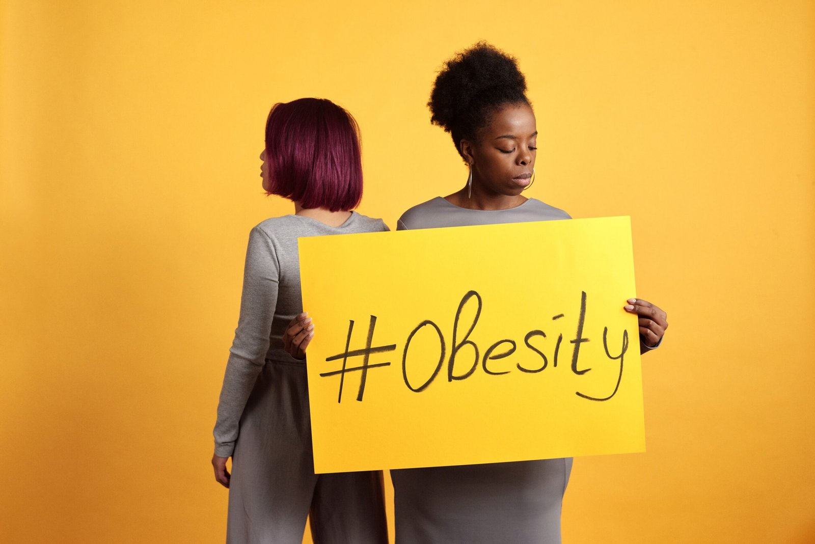 Two women advocate obesity awareness holding a bold sign with a vibrant yellow backdrop.