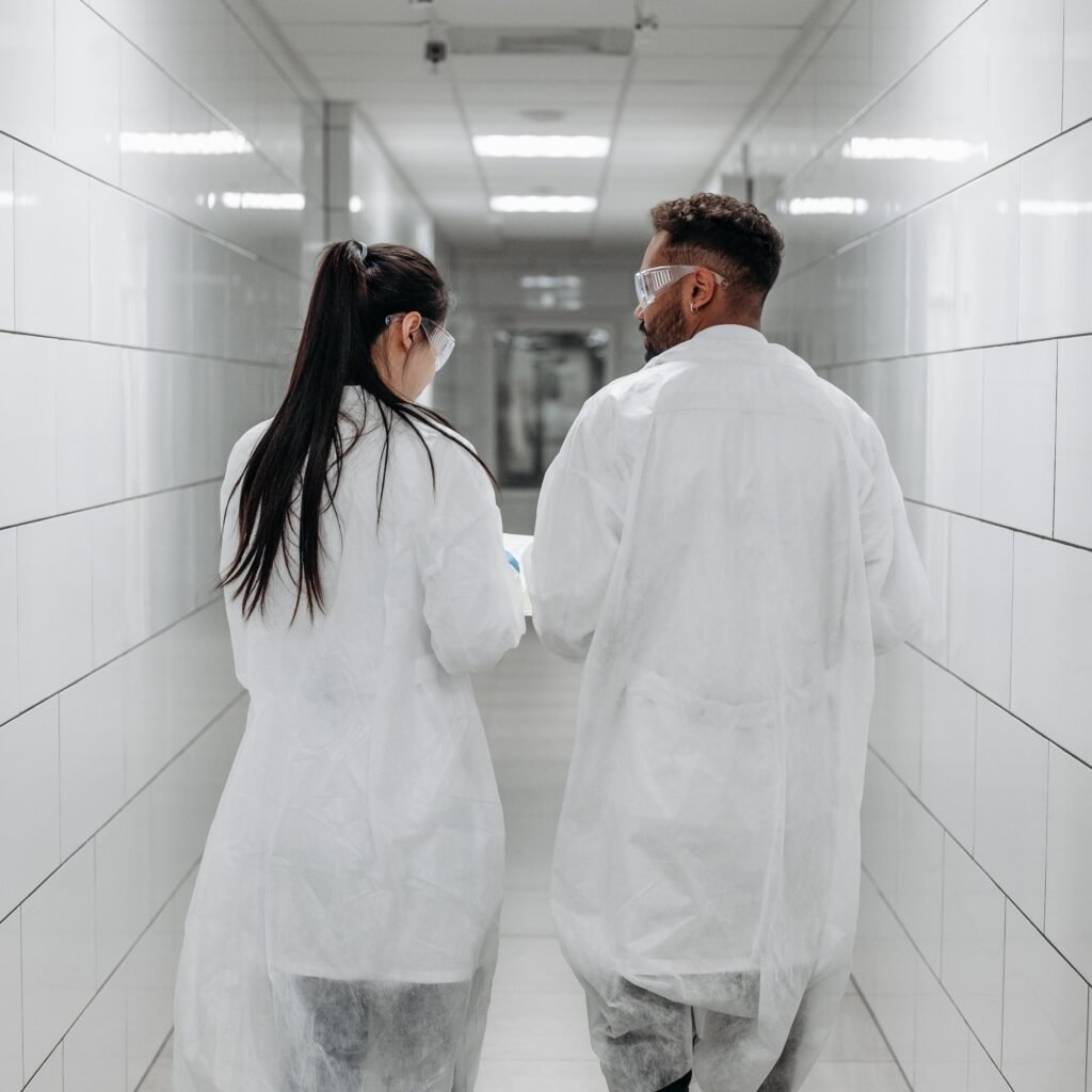 Two laboratory technicians in lab coats and goggles walking down a white tiled corridor, demonstrating teamwork and safety protocols.