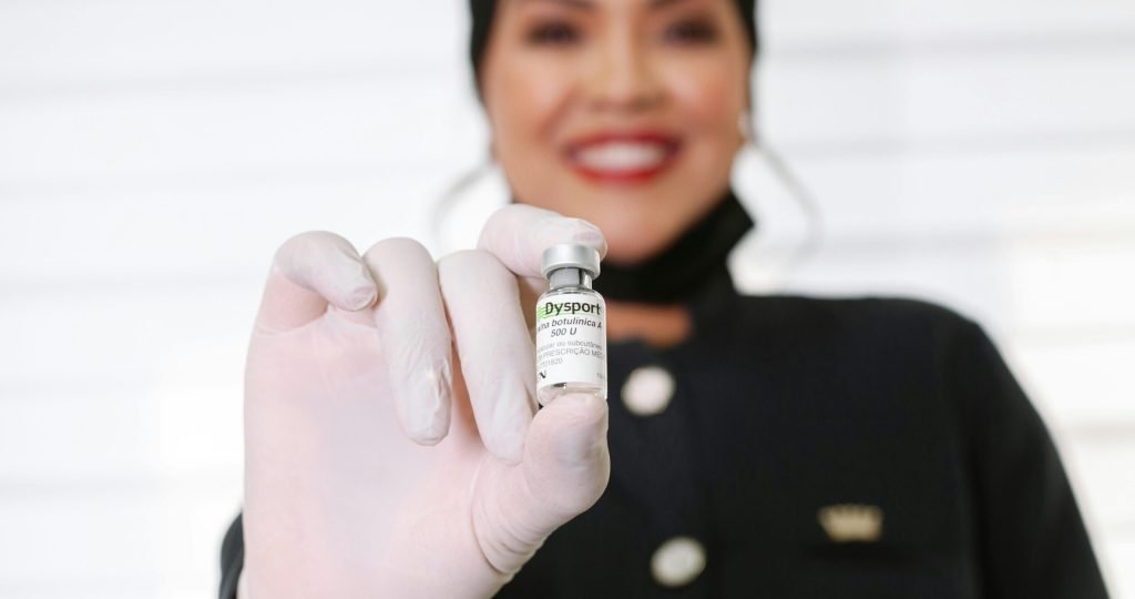 A healthcare worker in gloves holding a Dysport vial, smiling indoors.