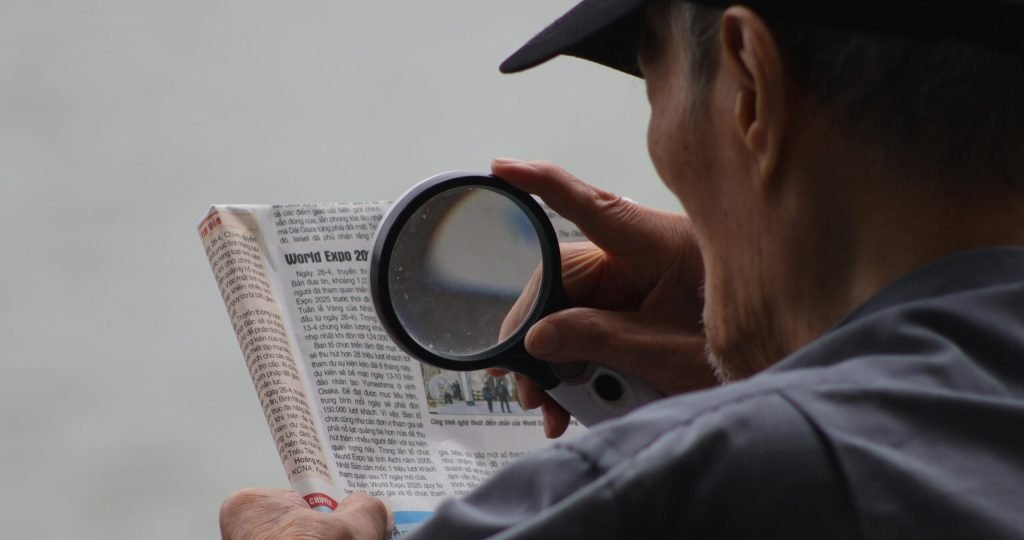 Elderly man using a magnifying glass to enhance text in a newspaper.