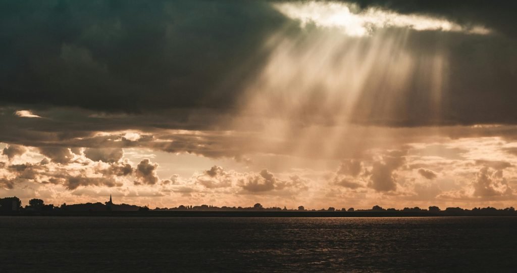 Sunbeams illuminate a serene landscape beneath dark storm clouds over a calm sea.