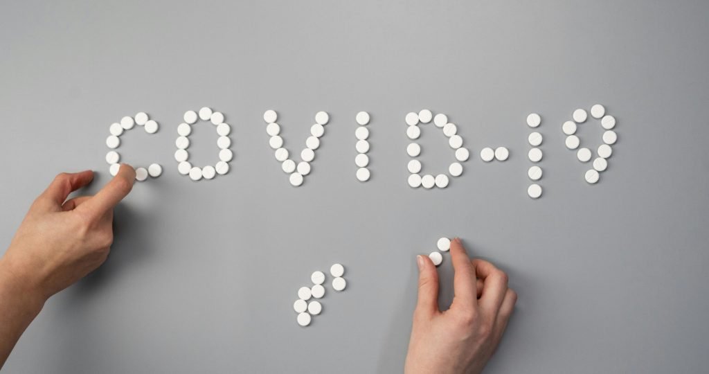 Hands arranging white pills to spell 'COVID-19' on a gray surface, symbolizing medical and health themes.