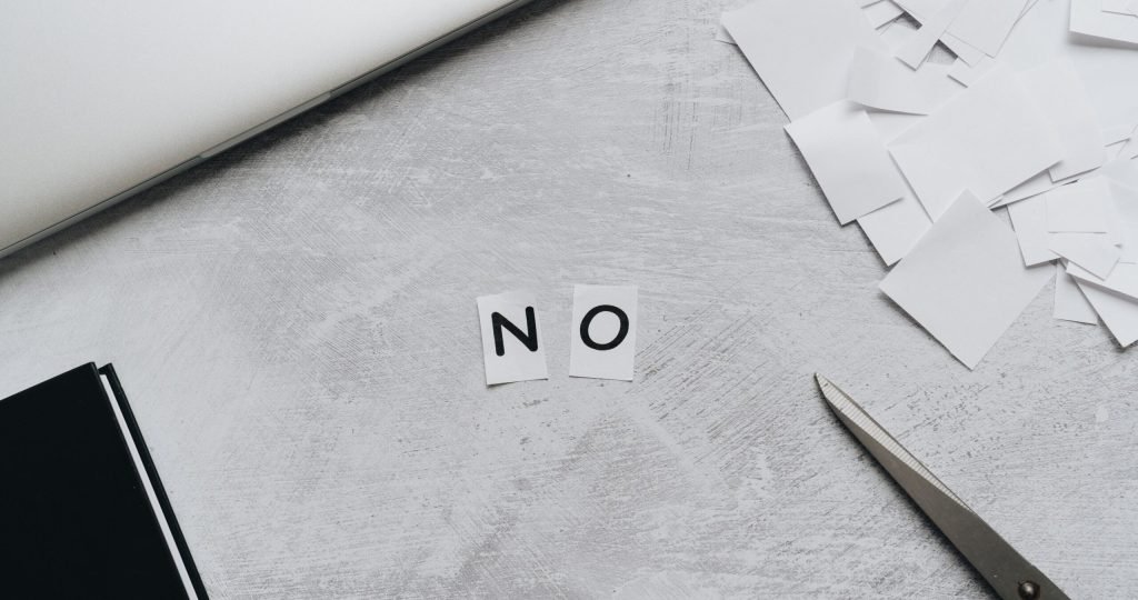 Top view of scattered paper squares, laptop, and scissors forming the word 'NO', implying rejection or denial.