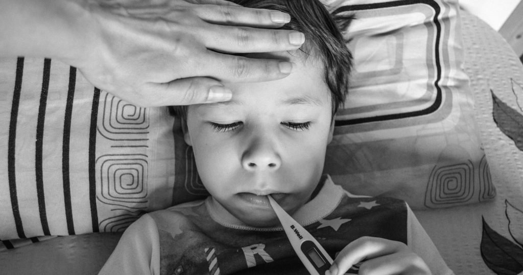 Black and white from above of unrecognizable person touching forehead of unemotional child in casual clothing holding electronic thermometer in mouth