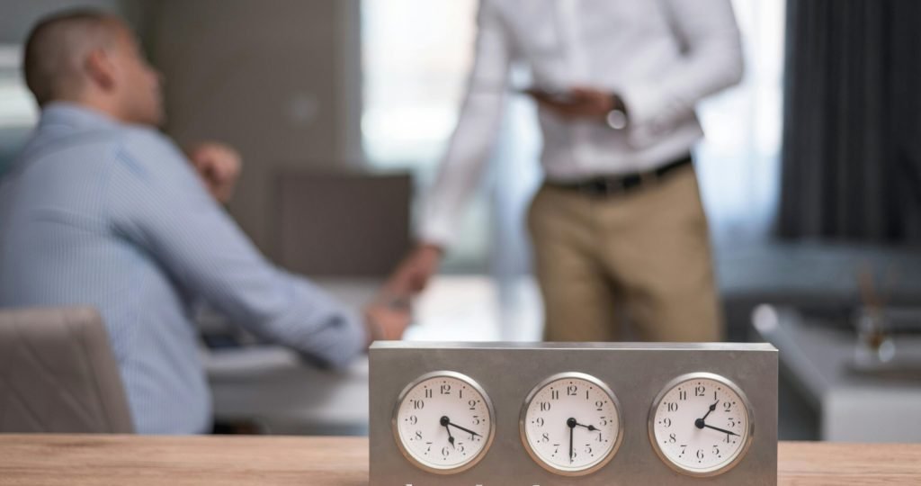 Two businessmen in an office with clocks showing different time zones, symbolizing global business.