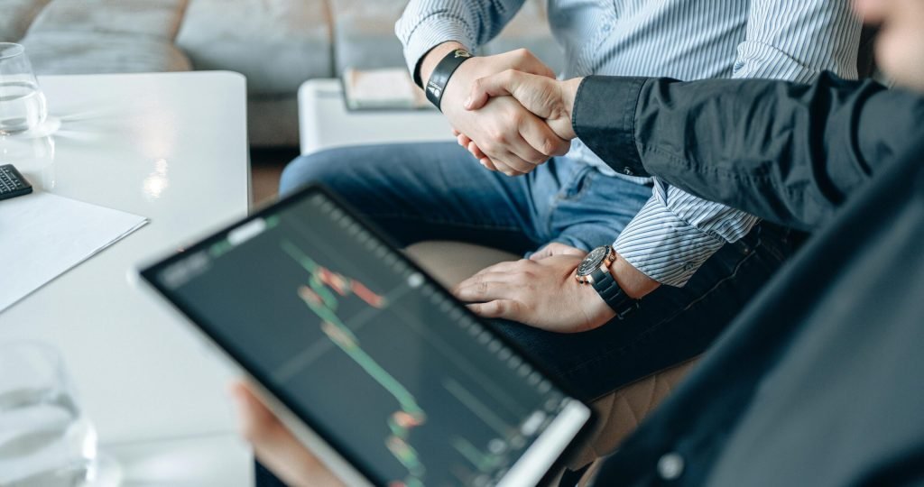Two businesspeople shaking hands over stock market data on a tablet.