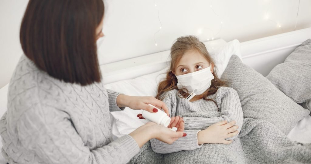 A mother helps her sick daughter wearing a face mask in bed, offering medicine.