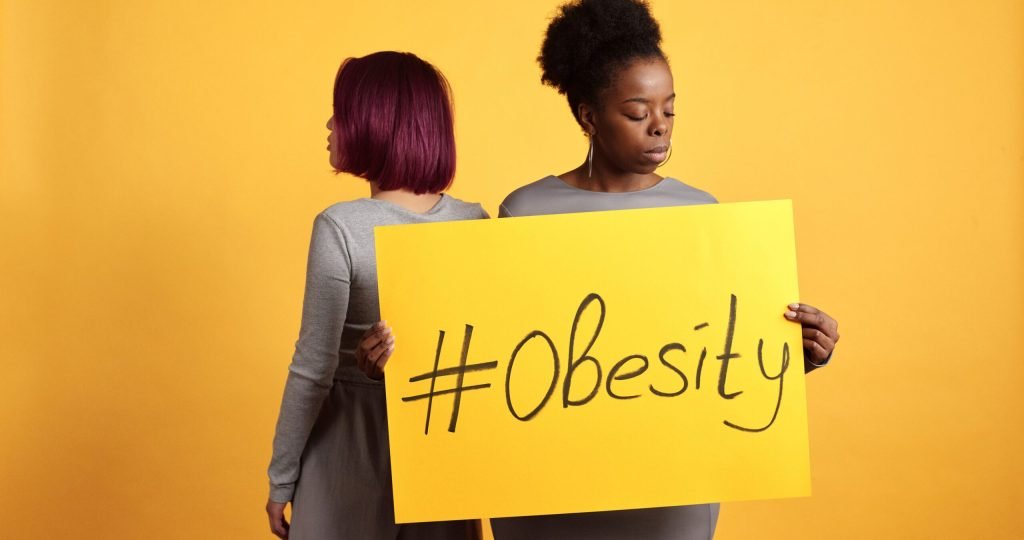 Two women advocate obesity awareness holding a bold sign with a vibrant yellow backdrop.