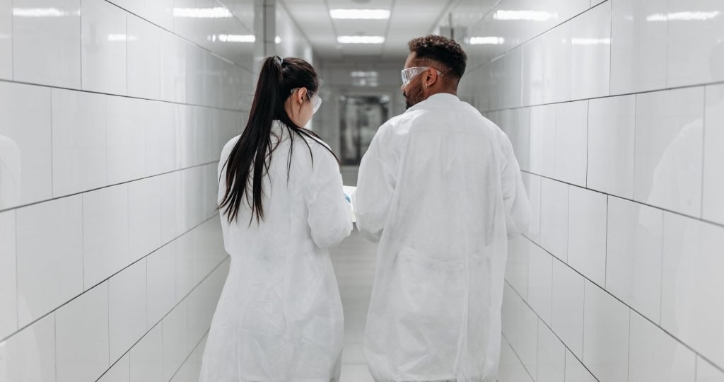 Two laboratory technicians in lab coats and goggles walking down a white tiled corridor, demonstrating teamwork and safety protocols.