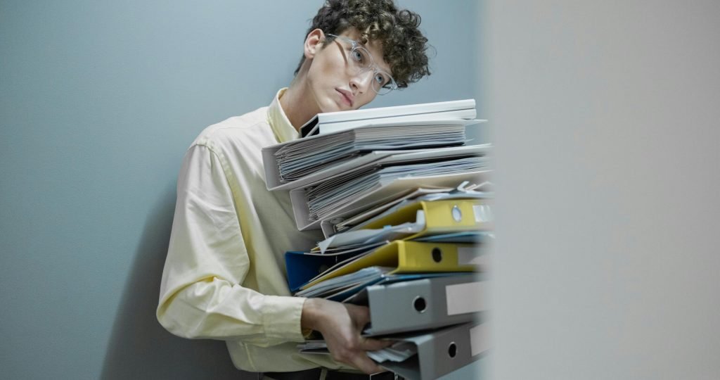 Young man overwhelmed with a stack of folders symbolizes workplace stress.