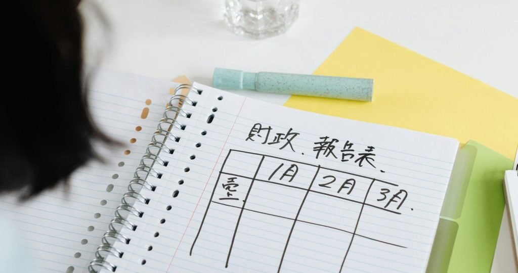 Person writing a table on a spiral notebook with planner and stationery on a desk.